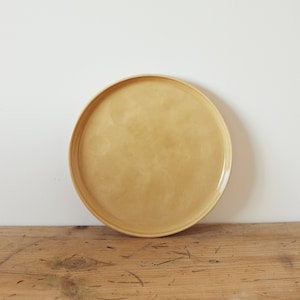 Può includere: Un piatto rotondo in ceramica di colore giallo chiaro con un bordo leggermente rialzato. Il piatto è appoggiato su una superficie di legno.