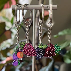 May include: Four keyrings with fruit designs, displayed on a silver stand. The keyrings feature purple grapes, pink raspberries, and blackberries. The stand is on a wooden surface. The keyrings are attached to silver chains.