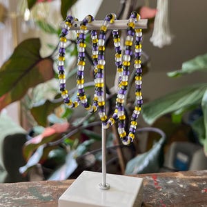 Pode incluir: Várias pulseiras de contas roxas, amarelas e transparentes exibidas em um suporte branco. O suporte está sobre uma superfície de madeira desgastada. Plantas verdes ao fundo.