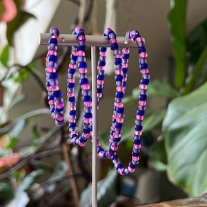 May include: A display of beaded bracelets in blue and pink hues. The bracelets are made of small, round beads. They are presented on a white stand, set against a backdrop of green foliage and a wooden surface.