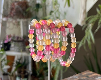 May include: A set of six colourful beaded bracelets stacked on a white display stand. The bracelets are made of translucent beads in shades of pink, yellow, and white.