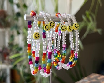 May include: A set of colourful beaded bracelets with smiley face charms. The bracelets are displayed on a white stand.