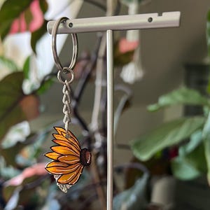 Op de afbeelding: Een zonnebloem sleutelhanger met een zilveren sleutelring en ketting. De zonnebloem heeft gele en oranje bloemblaadjes met een bruin midden. De sleutelhanger wordt getoond op een witte standaard tegen een achtergrond van groene planten.
