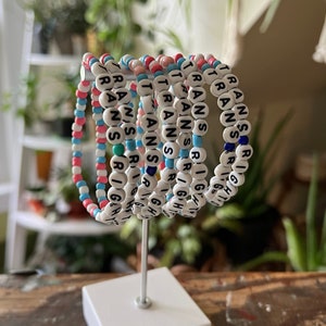 Peut inclure: Un ensemble de sept bracelets en perles avec les mots "TRANS RIGHTS" écrits en noir sur des perles blanches. Les bracelets sont empilés sur un support blanc et sont faits de perles roses, bleues et blanches.