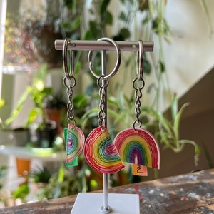 May include: Three rainbow-coloured keychains with silver chains and rings. The keychains are shaped like rainbows and have a variety of colours, including red, orange, yellow, green, blue, indigo, and violet. One keychain has the word "Love" written on it.