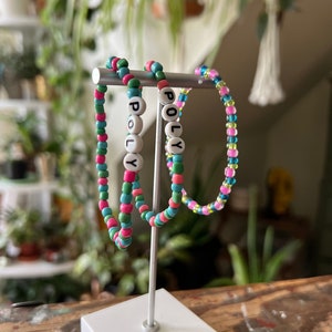 Peut inclure: Trois bracelets en perles colorées avec le mot "POLY" écrit en perles blanches. Les bracelets sont exposés sur un support blanc.