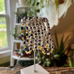 Könnte beinhalten: Ein Satz von acht Perlenarmbändern mit den Worten "they/them", die auf jedem Armband wiederholt werden. Die Armbänder sind auf einem weißen Ständer gestapelt und bestehen aus lila, gelben und weißen Perlen.