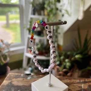 Puede incluir: Un brazalete de cuentas blancas con la palabra "PROTECT" escrita en letras negras. Las cuentas son de colores arcoíris y hay una pequeña cuenta en forma de corazón.