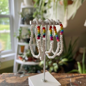 Puede incluir: Cinco pulseras de cuentas blancas con cuentas de colores arcoíris en un soporte blanco. Las pulseras están dispuestas en semicírculo.