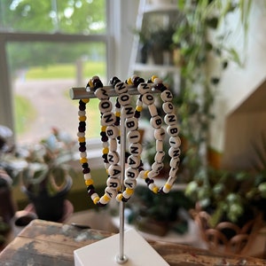 May include: Four beaded bracelets with the word "NONBINARY" spelled out in black letters on white beads. The bracelets are strung on a silver stand and have alternating black and yellow beads.