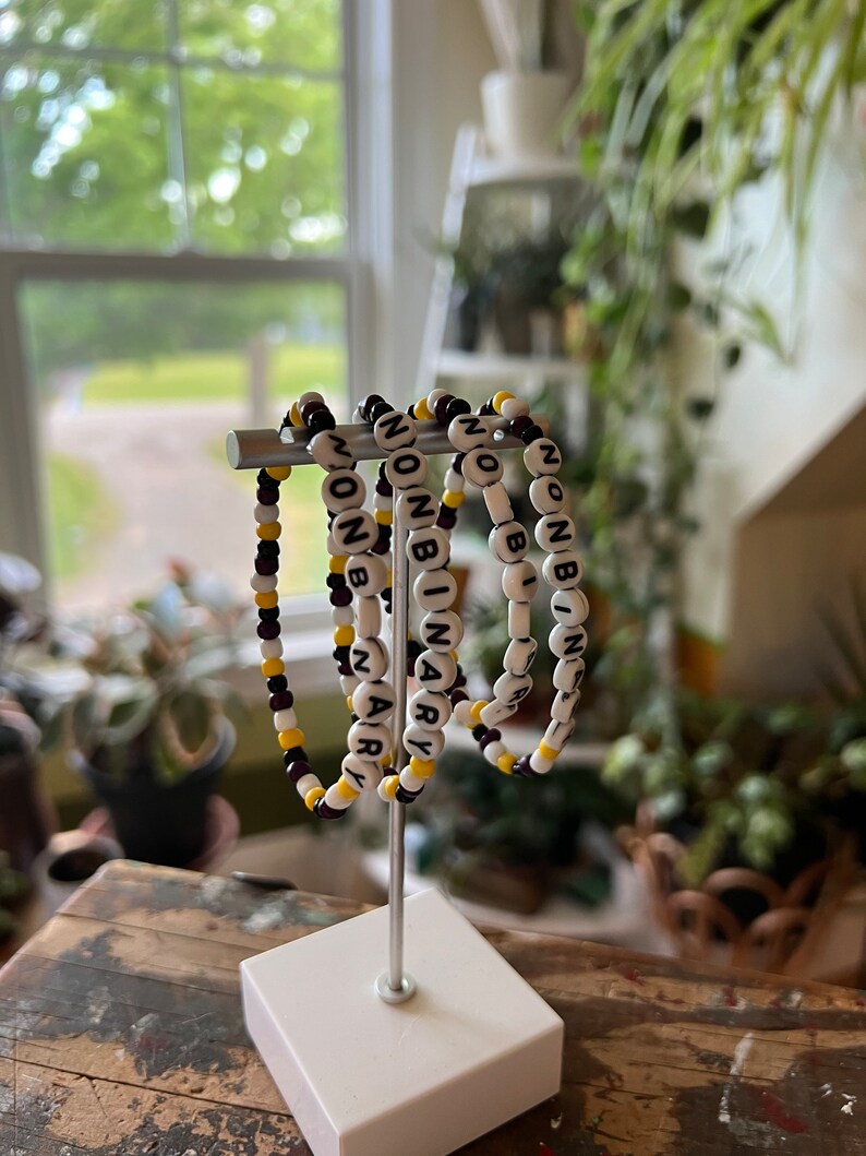 May include: Four beaded bracelets with the word "NONBINARY" spelled out in black letters on white beads. The bracelets are strung on a silver stand and have alternating black and yellow beads.