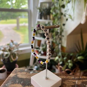 May include: A beaded bracelet with the word "NONBINARY" spelled out in white beads on a black background. The bracelet is made with purple, yellow, and white beads and is displayed on a white stand.