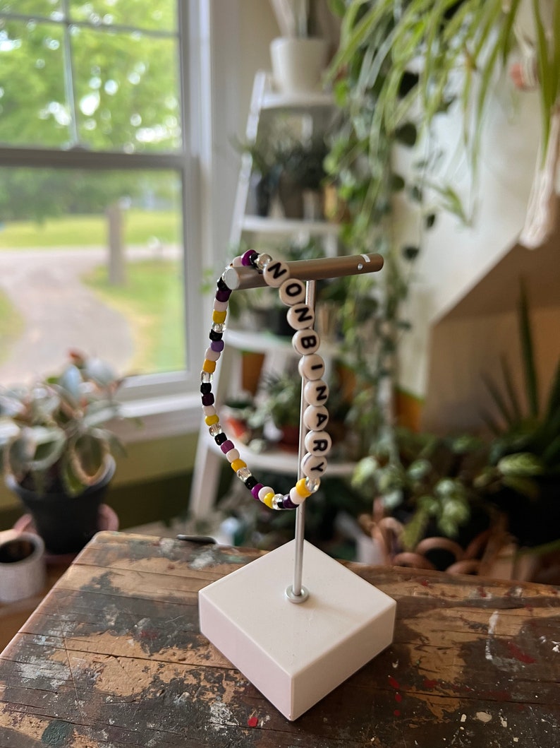 May include: A beaded bracelet with the word "NONBINARY" spelled out in white beads on a black background. The bracelet is made with purple, yellow, and white beads and is displayed on a white stand.