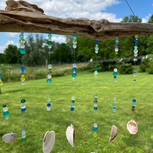 Peut inclure: Un carillon en bois flotté avec plusieurs brins de perles de verre vertes, bleues et transparentes et des coquillages blancs qui pendent. Le carillon est suspendu à l'extérieur sur fond d'herbe verte et de ciel bleu avec des nuages blancs.