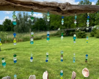 Puede incluir: Un carillón de madera flotante con múltiples hilos de cuentas de vidrio verde, azul y transparente y conchas blancas colgando de él. El carillón está colgado al aire libre sobre un fondo de hierba verde y cielo azul con nubes blancas.