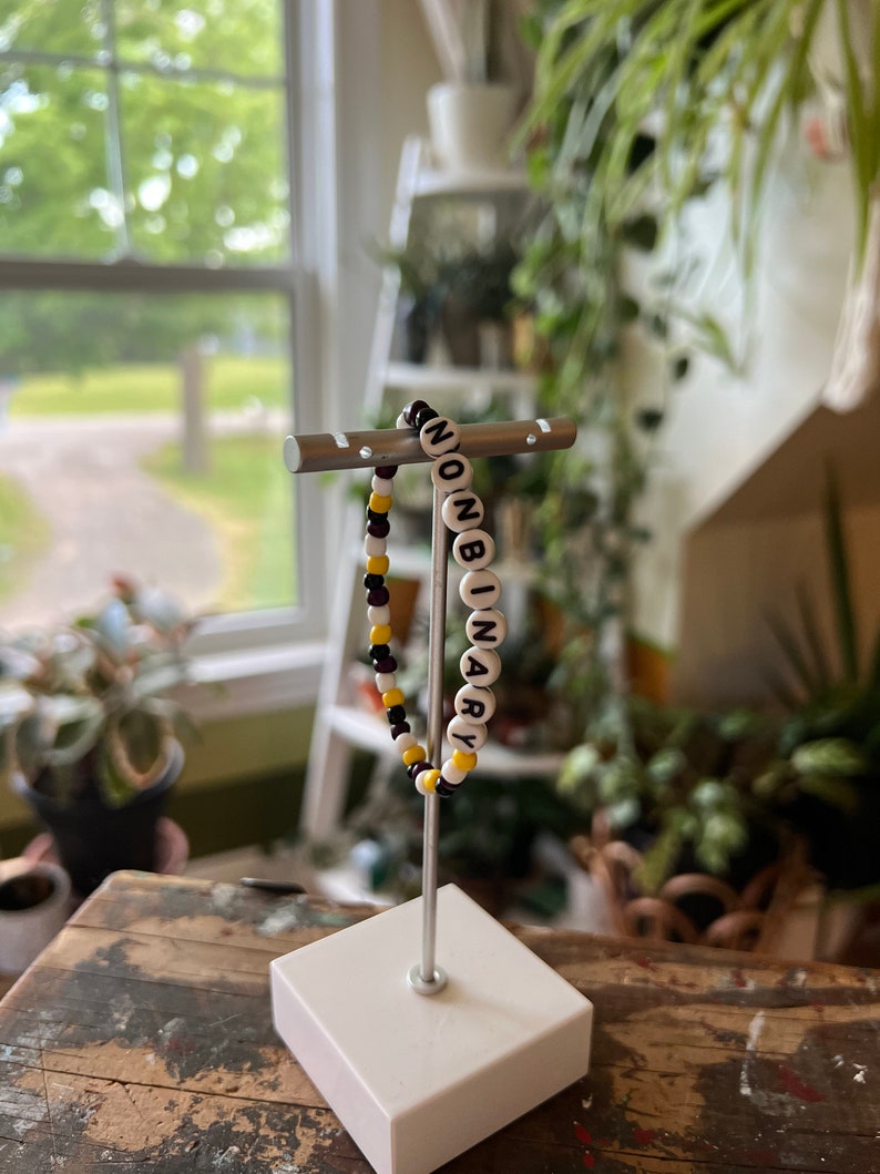 May include: A beaded bracelet with black and yellow beads spelling out the word "NONBINARY" in white letters.