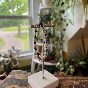 May include: A white beaded bracelet with black letter beads spelling "NON-BINARY". The bracelet is displayed on a silver stand with a white base.