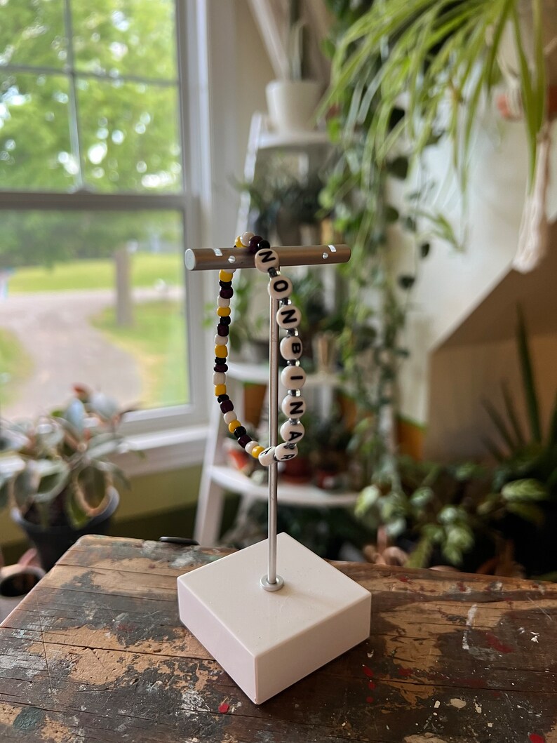 May include: A white beaded bracelet with black letter beads spelling "NON-BINARY". The bracelet is displayed on a silver stand with a white base.