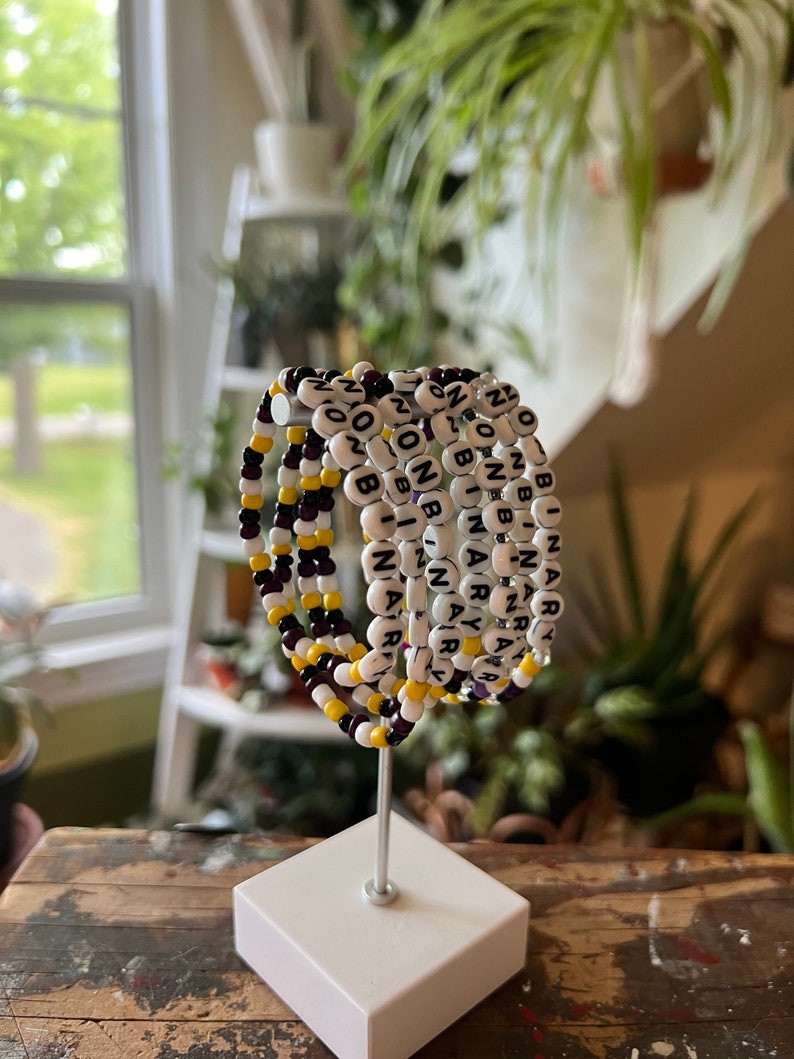 May include: A set of six beaded bracelets with the word "NARY" spelled out in black beads on a white background. The bracelets are stacked on a white stand and have alternating purple and yellow beads.