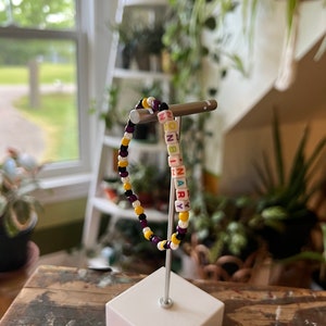 May include: A beaded bracelet with the word "NONBINARY" spelled out in colourful letter beads. The bracelet is made with purple, yellow and white beads.