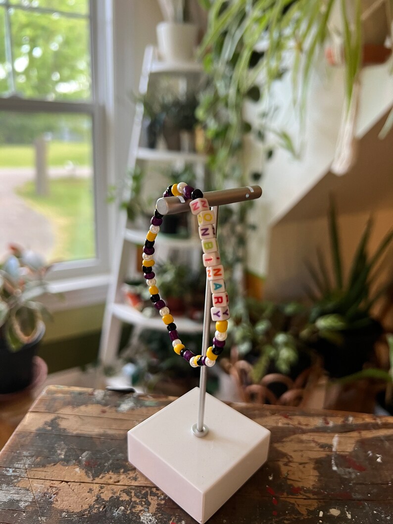 May include: A beaded bracelet with the word "NONBINARY" spelled out in colourful letter beads. The bracelet is made with purple, yellow and white beads.