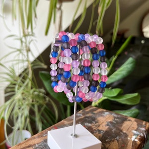 May include: A set of six colourful bracelets made of frosted glass beads. The bracelets are stacked on a white display stand. The beads are in shades of pink, purple, blue, and grey.
