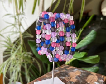 May include: A set of six colourful bracelets made of frosted glass beads. The bracelets are stacked on a white display stand. The beads are in shades of pink, purple, blue, and grey.