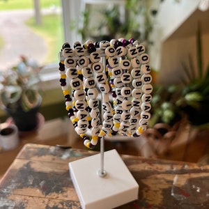 May include: A set of seven beaded bracelets with the words "nonbinary" spelled out in black beads on white beads. The bracelets are arranged in a circle on a white stand. The beads are interspersed with purple and yellow beads.
