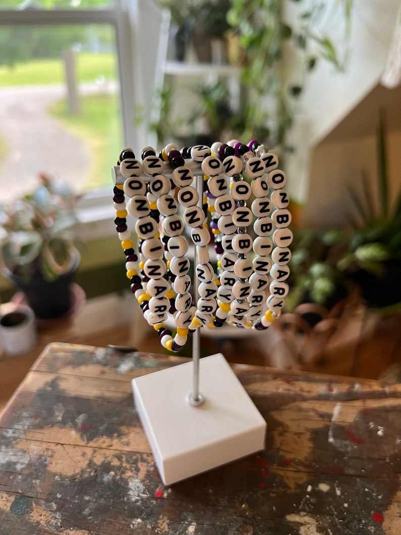 May include: A set of seven beaded bracelets with the words "nonbinary" spelled out in black beads on white beads. The bracelets are arranged in a circle on a white stand. The beads are interspersed with purple and yellow beads.