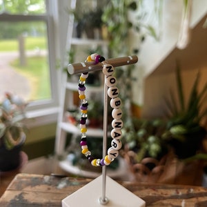 May include: A white beaded bracelet with the word "NONBINARY" spelled out in black letters. The bracelet is on a white stand and has yellow, purple, and black beads.