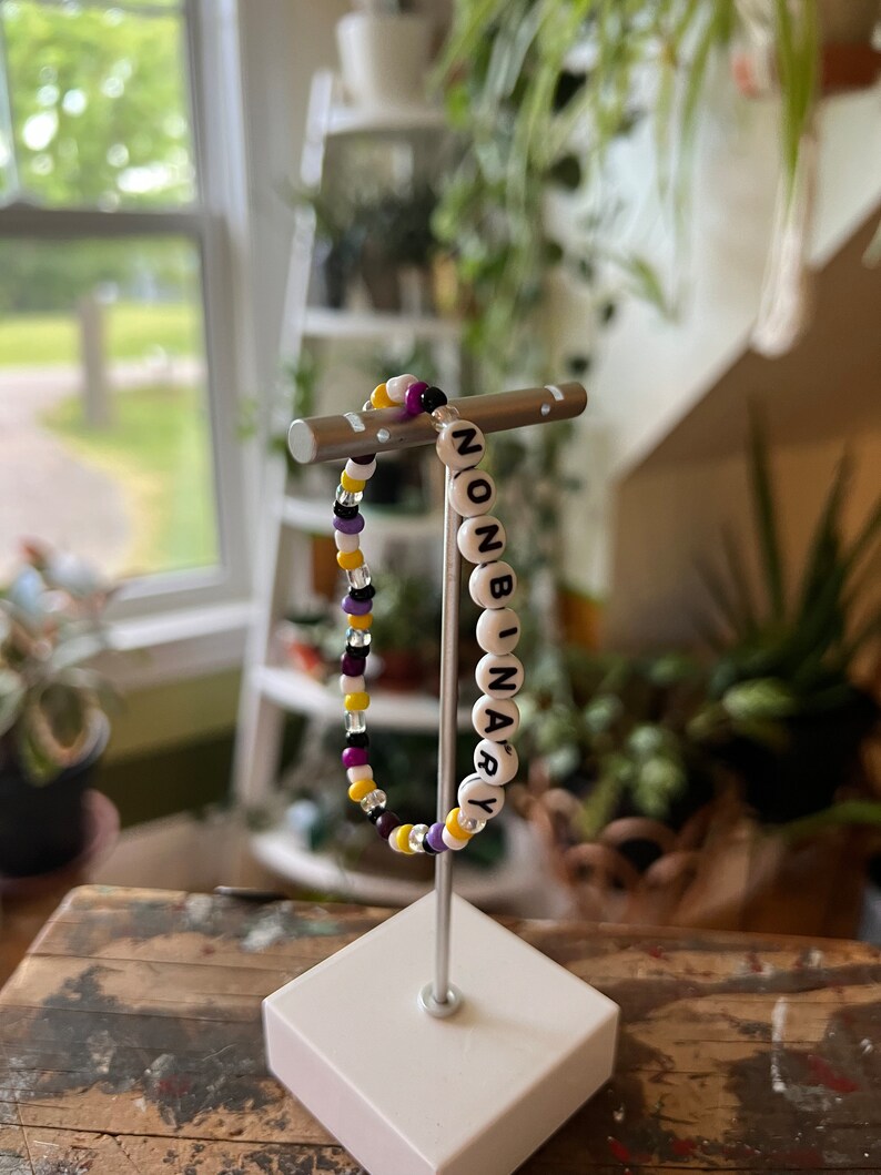 May include: A white beaded bracelet with the word "NONBINARY" spelled out in black letters. The bracelet is on a white stand and has yellow, purple, and black beads.