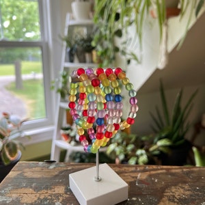 Peut inclure: Un ensemble de bracelets en perles colorées arc-en-ciel empilés sur un support blanc. Les bracelets sont faits de perles translucides dans différentes nuances de rouge, orange, jaune, vert, bleu et violet.