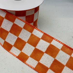 May include: A spool of wired ribbon featuring a repeating orange and white checkered pattern. The fabric has a textured weave, and the edges are finished with a matching orange stitched border, providing structure to the ribbon as it unrolls.