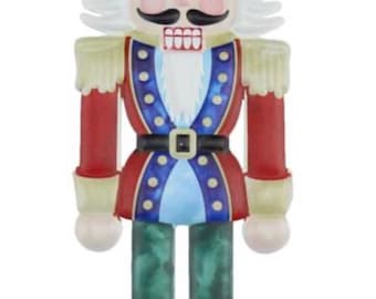 Puede incluir: Figura decorativa de cascanueces con un diseño de soldado clásico. La figura presenta un sombrero negro, cabello y bigote blancos, y un uniforme rojo y azul con detalles dorados. El cascanueces se apoya en botas negras con detalles de punto de cruz blancos.