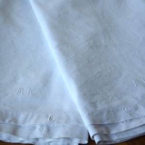 Op de afbeelding: Twee witte linnen handdoeken met geborduurde initialen "MK". De handdoeken zijn gevouwen en hebben een textuur. De onderste randen hebben kleine knoopdetails. De handdoeken liggen op een houten oppervlak.