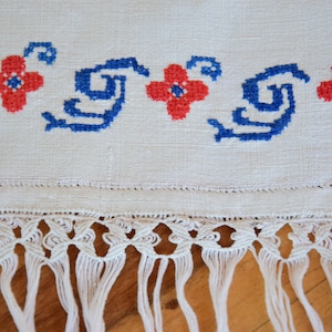 May include: White linen tea towel with a blue and red embroidered floral design and a white fringe border.