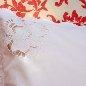 May include: White pillowcase with a delicate floral lace design. The lace is cut out, creating a see-through effect. The pillowcase is on a red and white floral patterned fabric.
