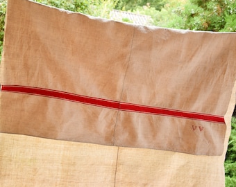 May include: A large, off-white linen fabric with a red stripe running horizontally across the center. The fabric has a rough, textured surface and appears to be old and worn. There are two small red triangles in the bottom right corner.