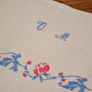 May include: A white linen tablecloth with blue and pink embroidered flowers and the letters "U" and "M" in blue.