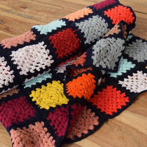 May include: A colorful, handmade crochet blanket with a dark blue border. The blanket features square patterns in various colors, including pink, white, red, orange, yellow, and gray. The blanket is folded and resting on a wooden surface.