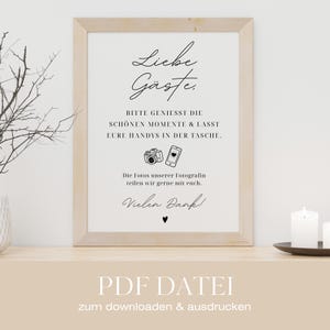 Puede incluir: Un letrero enmarcado con las palabras "Liebe Gäste" en escritura elegante. El letrero incluye texto en alemán, junto con ilustraciones de una cámara y un teléfono inteligente. El letrero está sobre una superficie de madera.