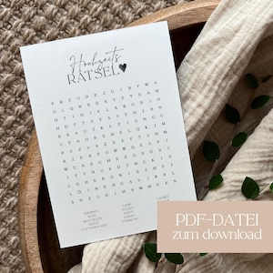 Op de afbeelding: Een witte kaart met de tekst "Hochzeits RÄTSEL" en een hartsymbool, met een woordzoekpuzzel. De kaart ligt in een houten kom met een beige doek en groene bladeren. De rechter benedenhoek heeft de tekst "PDF-DATEI zum download".