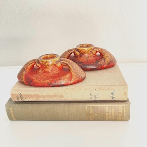 May include: Two ceramic, reddish-orange vases with a textured surface and a unique shape, featuring a central opening and curved handles. The vases are displayed on top of two vintage books with aged covers, creating a warm, earthy aesthetic.