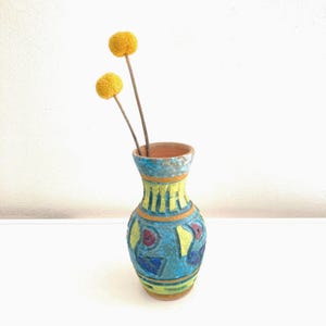 May include: A hand-painted ceramic vase with a blue, yellow, and orange geometric design. The vase holds two yellow, spherical flowers on brown stems. The vase has a textured, rustic appearance and is set against a white background.