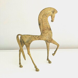 May include: A gold-colored, stylized horse sculpture with an elongated neck and raised front leg. The horse has a slender body and detailed texture, suggesting an antique or vintage aesthetic. The sculpture is set against a plain white background.