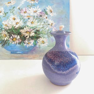 May include: A small, lavender-colored ceramic oil lamp with a speckled design and a wick. The lamp has a rounded body and a slender neck. A painting of white daisies in a blue vase is in the background.