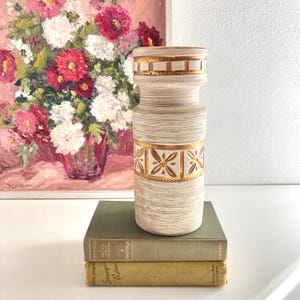 May include: White ceramic vase with gold accents and a floral pattern. The vase is sitting on top of two books.
