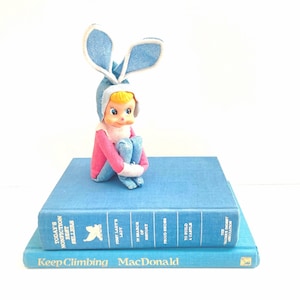 May include: A vintage doll wearing a blue bunny hat and pink outfit sits atop two blue books. The doll has blonde hair and is in a seated position. The books have titles such as "Today's Nonfiction Best Sellers" and "Keep Climbing MacDonald."