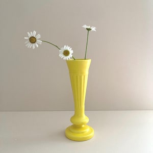 May include: A bright yellow ceramic vase with a flared rim, a slender neck featuring vertical fluted detailing, and a wide circular base. Two white daisies with yellow centers are placed inside the vase, extending outward from the opening.