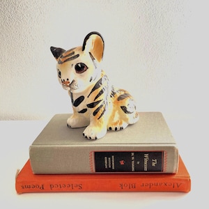 May include: A ceramic tiger cub figurine with black stripes and spots, perched atop two vintage books. The top book is gray with a black and red label, while the bottom book is orange with gold lettering. The figurine has a cream-colored body and large, dark eyes.